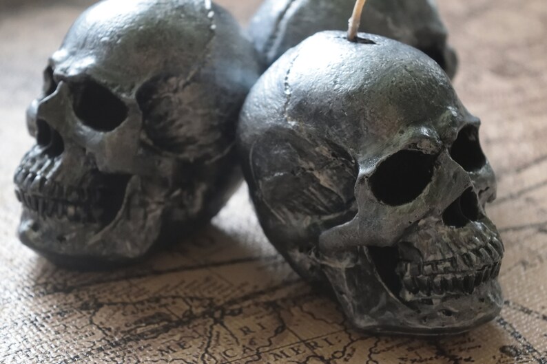 May include: Close-up of three gray skull-shaped candles. The candles have a textured, metallic finish and are arranged on a map-like surface. The candles are a decorative item, perfect for Halloween or gothic decor.