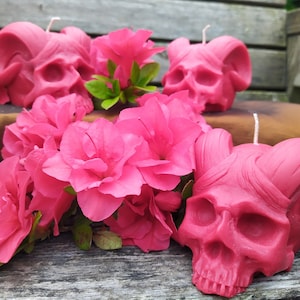 Puede incluir: Tres velas de calavera rosas con cuernos, rodeadas de flores de azalea rosas. Las velas son de cera y tienen un diseño de calavera detallado.