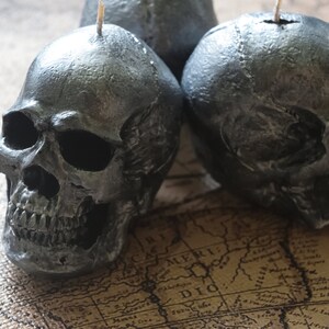 May include: Three dark gray skull-shaped candles with visible details of the skull's structure. Each candle has a wick at the top. The candles are placed on a vintage map, creating a gothic or Halloween theme.