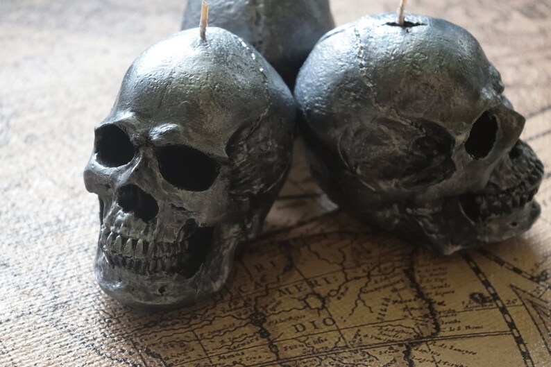 May include: Three dark gray skull-shaped candles with visible details of the skull's structure. Each candle has a wick at the top. The candles are placed on a vintage map, creating a gothic or Halloween theme.