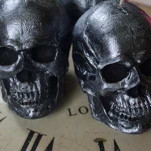 May include: Two metallic gray skull-shaped decorative objects. The skulls have detailed features, including eye sockets, nasal cavities, and teeth. The skulls are placed on a clock face, creating a gothic or Halloween theme.