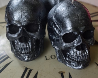 Può includere: Due oggetti decorativi a forma di teschio grigio metallizzato. I teschi presentano dettagli, tra cui orbite oculari, cavità nasali e denti. I teschi sono posti su un quadrante di orologio, creando un tema gotico o di Halloween.