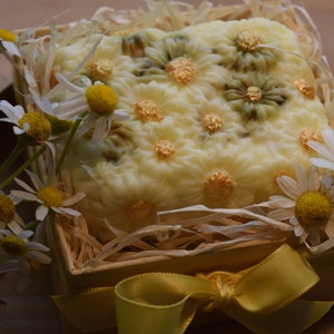 May include: A white soap bar with a floral design and gold accents is displayed in a wooden box with straw and a yellow ribbon. The box is surrounded by white daisies.
