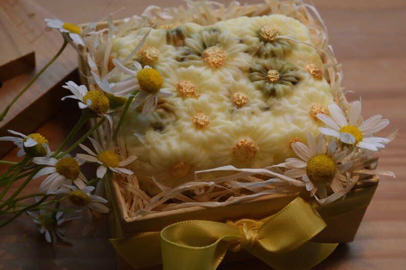 May include: A white soap bar with a floral design and gold accents is displayed in a wooden box with straw and a yellow ribbon. The box is surrounded by white daisies.