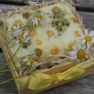 May include: A white and yellow handmade soap bar with a floral design, decorated with gold accents, in a small wooden box with a yellow ribbon. The box is filled with shredded paper and surrounded by white daisies.