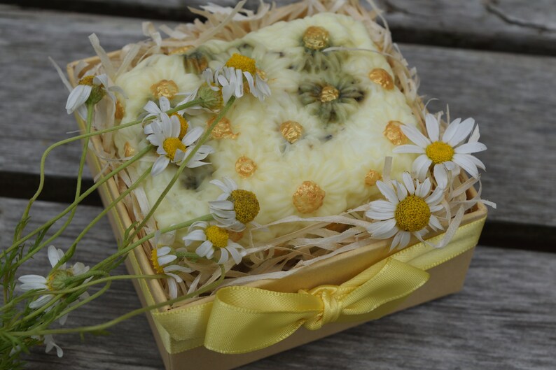 May include: A white and yellow handmade soap bar with a floral design, decorated with gold accents, in a small wooden box with a yellow ribbon. The box is filled with shredded paper and surrounded by white daisies.