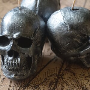 May include: Three silver skull-shaped candles with wicks, arranged on a vintage map. The candles have a metallic finish, and the skulls are detailed with facial features. The background is a sepia-toned map.