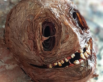May include: A brown, textured, paper mache mask with a wide, toothy grin. The mask has two large, dark eye holes and a slightly sinister expression.