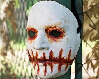 May include: A white full-face mask featuring dark eye sockets surrounded by reddish-brown painted details and a mouth area depicted with vertical stitched lines in a similar reddish-brown color. The text Sadofekalus.com is visible.