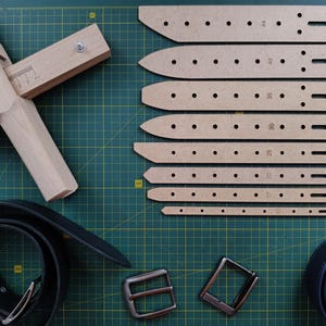 Op de afbeelding: Een set van acht bruine leren riem sjablonen met verschillende maten die erop zijn aangegeven. De sjablonen liggen op een groene snijmat. Er zijn ook twee zilveren gespen en een leren riem op de mat.