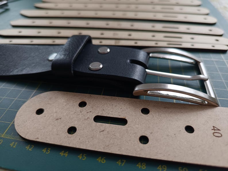 Op de afbeelding: Een zwarte leren riem met een zilveren gesp en een bruine leren band met gaten om de maat aan te passen. De band is genummerd van 40 tot 51.