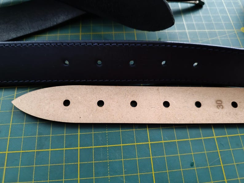 Op de afbeelding: Close-up van een donkerblauwe leren riem met meerdere stiknaden en vijf gelijkmatig verdeelde gaten. De riem ligt bovenop een lichtbruine leren riem met zeven gelijkmatig verdeelde gaten en het nummer "30" gedrukt aan de rechterkant.