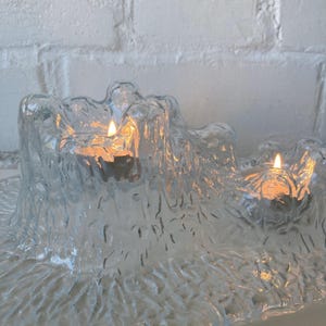 May include: A clear glass candle holder with a textured, organic design. The holder has three wells, each containing a lit candle with a warm, orange glow. The background is a white brick wall.