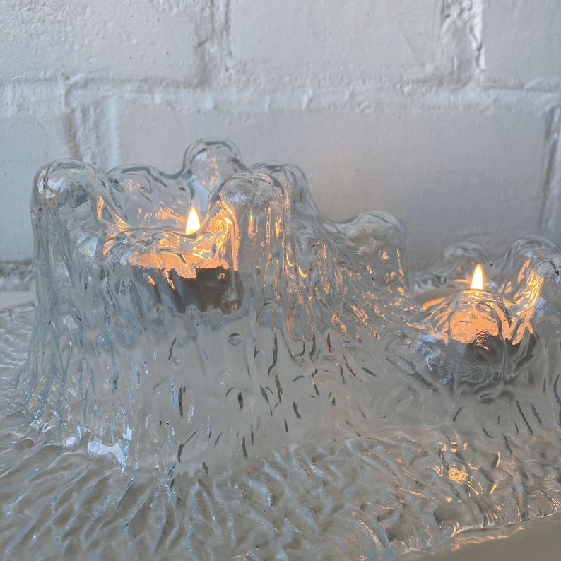 May include: A clear glass candle holder with a textured, organic design. The holder has three wells, each containing a lit candle with a warm, orange glow. The background is a white brick wall.