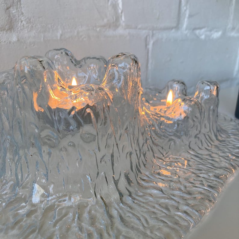 May include: A clear glass candle holder with a textured, organic design. The holder has multiple wells, each containing a lit candle. The flames cast a warm glow against the textured glass, creating a cozy ambiance.