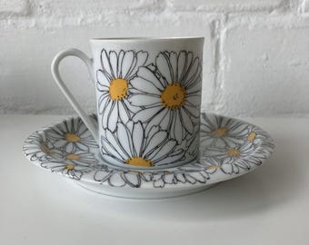 May include: White ceramic teacup and saucer set decorated with large daisy floral pattern. The daisies have white petals, black outlines, and yellow centers. The cup has a curved handle.