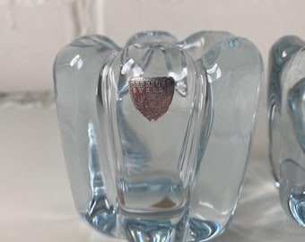 May include: Two light blue glass candle holders with a unique, petal-like design. Each holder has a clear, textured surface and a small, red and silver label. The candle holders are made of glass.