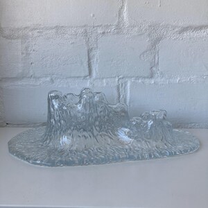 May include: A clear glass decorative piece with a textured, organic design. The piece has a wide, oval base with a raised, undulating form resembling a landscape or abstract sculpture. The glass has a frosted appearance.