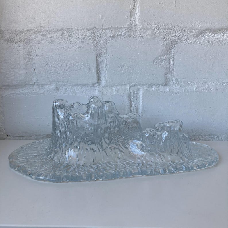 May include: A clear glass decorative piece with a textured, organic design. The piece has a wide, oval base with a raised, undulating form resembling a landscape or abstract sculpture. The glass has a frosted appearance.