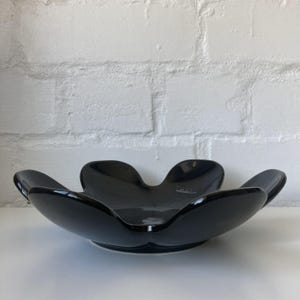 Könnte beinhalten: Eine glänzend schwarze Dekoschale mit einem einzigartigen, blütenblattartigen Design. Die Schale hat eine breite, offene Form und steht auf einer weißen Oberfläche. Der Hintergrund ist eine weiße Backsteinmauer.