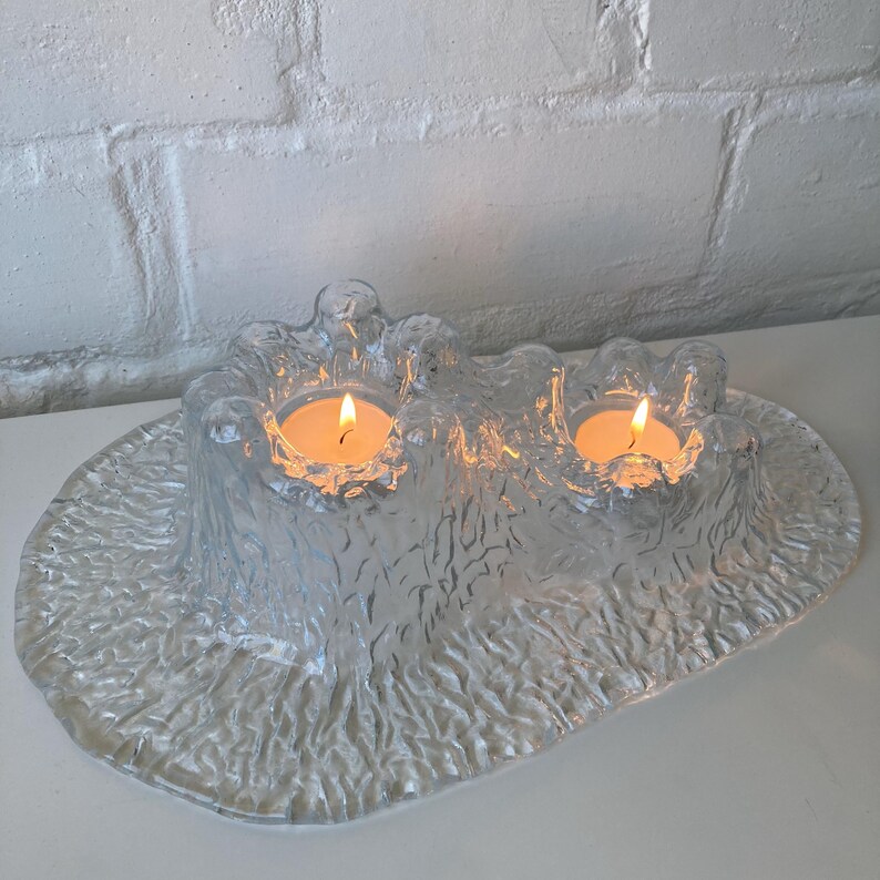May include: A clear glass candle holder with two lit candles. The holder has a textured, organic design and sits on a white surface. The flames of the candles are bright against the clear glass.