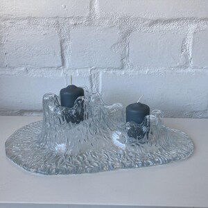 May include: A clear glass candle holder with a textured, organic design. The holder has two dark gray candles with white wicks. The base is oval-shaped, and the holder has a rough, uneven surface. The background is a white brick wall.