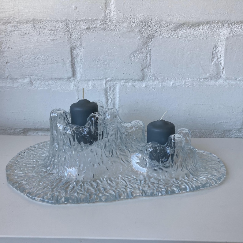 May include: A clear glass candle holder with a textured, organic design. The holder has two dark gray candles with white wicks. The base is oval-shaped, and the holder has a rough, uneven surface. The background is a white brick wall.