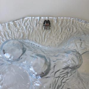 May include: A clear, textured glass bowl with a wavy design. The bowl has a rippled surface and a few rounded, bubble-like features. The brand "muurla hand made in finland" is printed on a label at the top of the bowl.