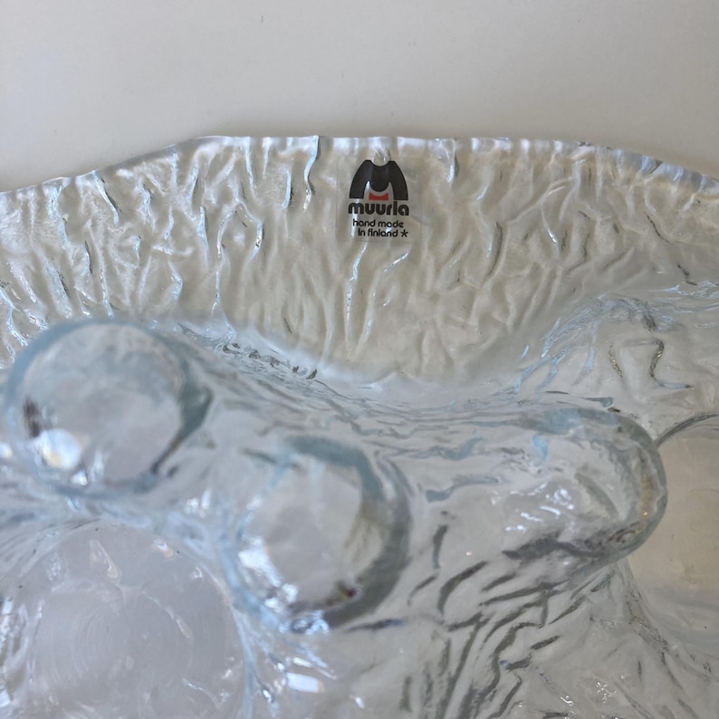May include: A clear, textured glass bowl with a wavy design. The bowl has a rippled surface and a few rounded, bubble-like features. The brand "muurla hand made in finland" is printed on a label at the top of the bowl.