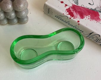 May include: A green glass candle holder with a unique, elongated shape. The holder has two circular indentations for candles and a curved design. A book and a gray bubble candle are in the background.