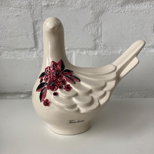 Puede incluir: Figura de paloma de cerámica color crema con diseño floral. La paloma tiene un acabado brillante y un racimo de flores rojas con hojas negras en un lado. La base de la paloma tiene el texto "Design Rosa Ljung".