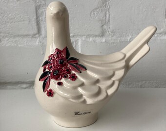 May include: A cream-colored ceramic dove figurine with a floral design. The dove has a smooth, glossy finish and a cluster of red flowers with black leaves on its side. The base of the dove has the text "Design Rosa Ljung".