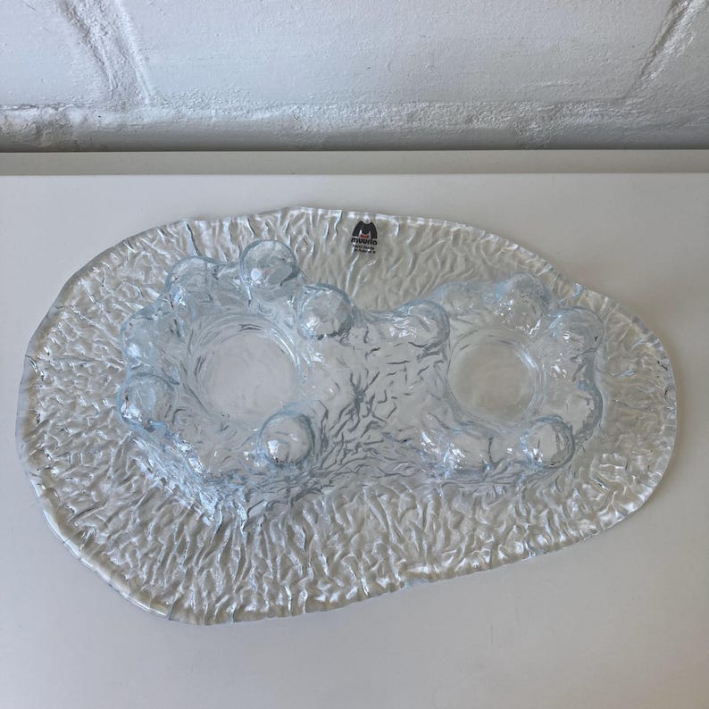 May include: A clear glass candle holder with a textured, organic design. The holder features two circular spaces for candles, surrounded by bubble-like shapes. The base has a textured, rippled surface. The brand logo is visible.
