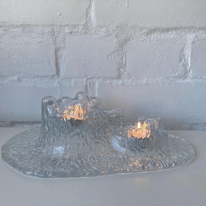 May include: A clear glass candle holder with a textured surface. The holder has a tiered design with two lit candles, creating a warm glow. The base is a wide, oval shape.