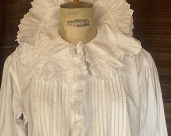 May include: A white cotton blouse with a large, ruffled collar and a row of vertical pleats down the front. The collar has a delicate lace trim.