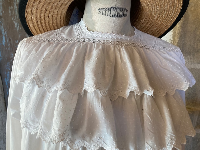 Op de afbeelding: Witte katoenen blouse met een gerimpelde halslijn en een gestippeld patroon. De blouse heeft een delicate kantversiering rond de halslijn en de ruches.