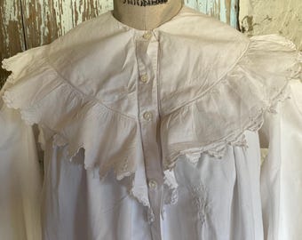 May include: White cotton blouse with a large, ruffled collar and embroidered details. The collar is trimmed with lace and the blouse has long sleeves.