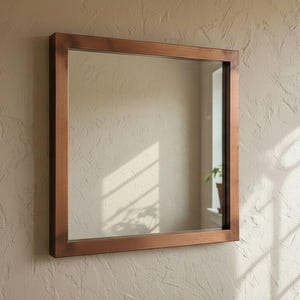 Puede incluir: Espejo cuadrado con marco de madera colgado en una pared beige texturizada. El espejo refleja un vistazo de una habitación con una planta en maceta. Un jarrón blanco con plantas secas descansa sobre una repisa de madera.