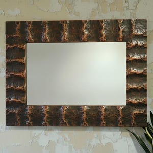 Puede incluir: Espejo rectangular con un marco texturizado de color cobre. El marco tiene un patrón repetitivo de formas rectangulares en relieve. El espejo está montado en una pared de color claro. Una planta verde es visible en la esquina inferior derecha.