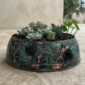 May include: A round, low-profile planter with a hammered copper finish featuring a dark green and black patina. The planter is filled with dark soil and contains a variety of small green succulents and a small leafy plant with rounded foliage.