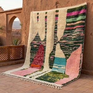 May include: A large, rectangular rug with a cream-colored background and colorful geometric designs. The rug features pink, green, and blue shapes, with a fringed edge. It is leaning against a textured, reddish-brown wall, suggesting an outdoor setting.