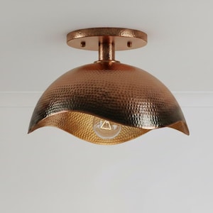 May include: A copper-colored ceiling light fixture with a hammered texture. The light has a dome-shaped shade with a wavy edge, and a gold-colored interior. The fixture is mounted on a white ceiling.
