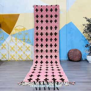 May include: A pink and brown Moroccan wool rug with a repeating cross pattern. The rug has a rectangular shape with a fringe on the bottom. The rug is displayed on a gray floor with a colorful background.