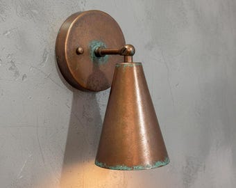 May include: A copper-colored wall sconce with a round backplate and a cone-shaped shade. The fixture has a vintage look with a slight patina, suggesting age and use. The sconce is mounted on a gray wall.