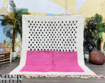 May include: A white and pink Moroccan wool rug with a black geometric pattern. The rug has a fringe border.