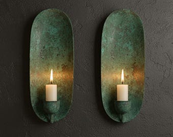 Puede incluir: Dos apliques de pared con acabado de pátina verde, cada uno con una vela blanca encendida. Los apliques tienen forma ovalada alargada y están montados en una pared gris oscura texturizada. Las llamas de las velas emiten un brillo cálido.