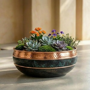 Puede incluir: Una maceta redonda de metal martillado con un acabado verde oxidado oscuro en la parte inferior y una banda de cobre pulido en el borde. El cuenco está lleno de una variedad de pequeñas suculentas, flores de color naranja brillante y pensamientos morados.