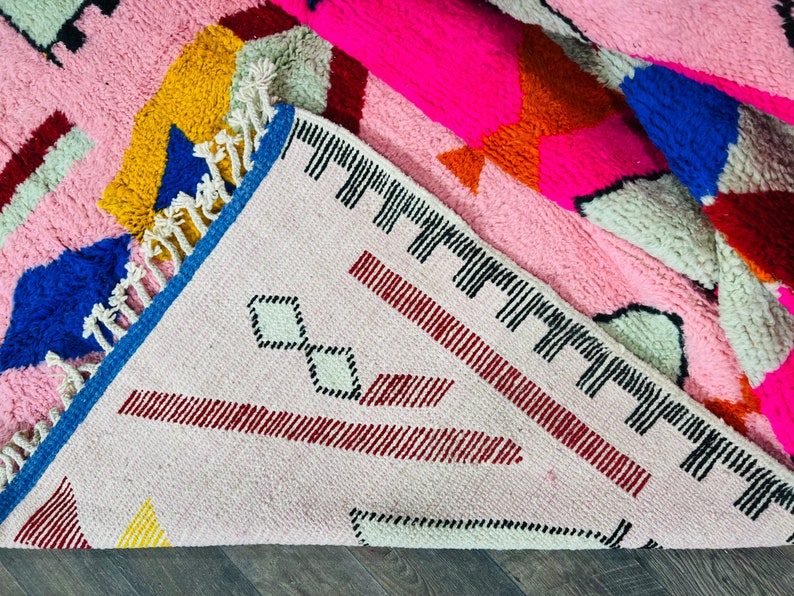May include: A pink, blue, yellow, and red geometric patterned rug with fringe. The rug has a white background with a black diamond pattern.