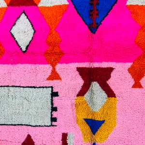 May include: A pink and white rug with a geometric pattern. The rug features a variety of colors, including red, orange, yellow, blue, and brown. The rug is made of a soft, plush material.