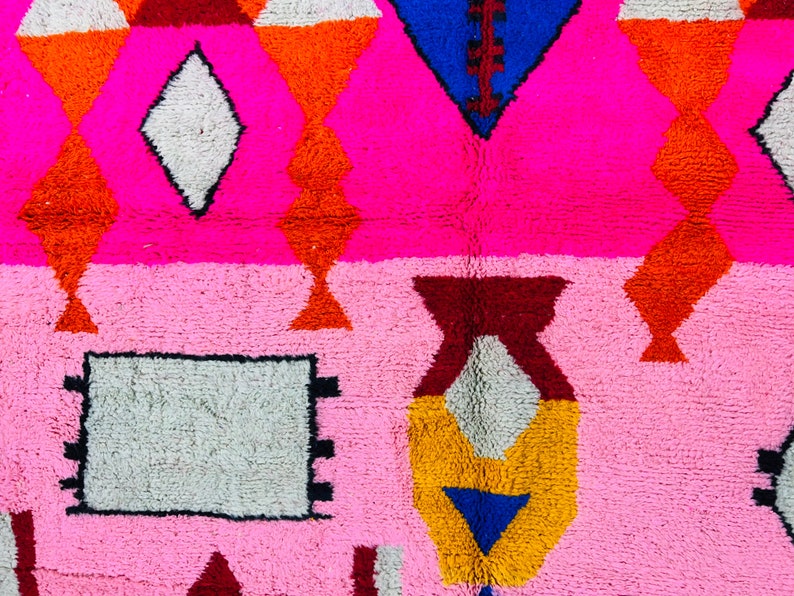 May include: A pink and white rug with a geometric pattern. The rug features a variety of colors, including red, orange, yellow, blue, and brown. The rug is made of a soft, plush material.